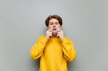 Hasta genç adam koruyucu maskesini çıkardı ve gri duvara doğru hapşırdı. Tıbbi maskeli hasta bir adam üşütmüş..