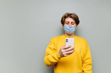 Koruyucu maskeli genç adam gri arka planda akıllı telefon kullanıyor, odaklanmış bir yüzle ekrana bakıyor..