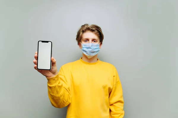 医療用マスクのハンサムな若い男は 灰色の壁の背景に白い画面を持つスマートフォンを示しています 男の手の中にスマートフォンに焦点を当てます 隔離されてる コピースペース — ストック写真