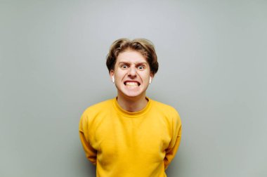 Gri arka planda kablosuz kulaklıklı öfkeli genç bir adamın portresi, kameraya bakıyor ve dişlerini öfkeyle gösteriyor. İzole edilmiş.