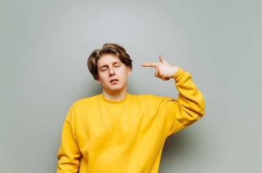 Üzgün genç bir adam, gri bir duvarın arka planında gözleri kapalı bir silahla kafasını vuruyor. İzole edilmiş.