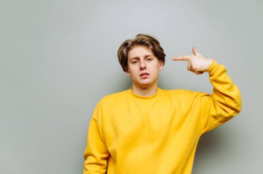 Sarı elbiseli yorgun genç adam kameraya bakar ve bir parmağını kafasından vurur ve yorgunluktan intihar etmek ister. Gri arkaplanda izole.