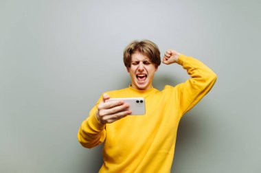 Elinde akıllı telefon olan neşeli genç adam zaferle seviniyor. Elleri havada, bağırıyor ve gri bir duvarın arka planındaki telefon ekranına bakıyor..