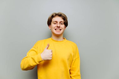 Sarı kazaklı olumlu bir genç parmak hareketi gösteriyor ve yüzünde bir gülümsemeyle kameraya bakıyor. İzole edilmiş