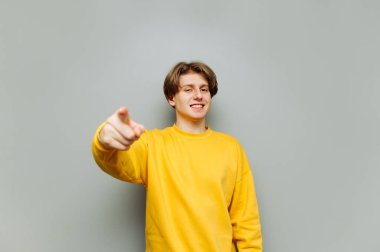 Mutlu genç adam gri bir arka planda duruyor ve yüzünde bir gülümsemeyle kameraya bakıyor ve parmağını gösteriyor. İzole edilmiş.