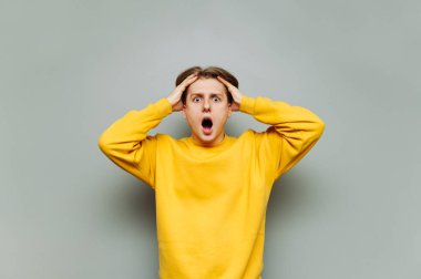 Gri bir duvarın arka planında parlak gündelik giysiler içinde haykırışlarla kameraya bakan ve kafasıyla el ele tutuşan şaşırmış genç bir adamın portresi..