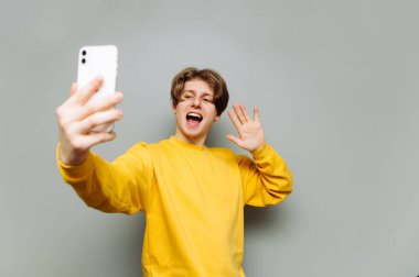 Sarı tişörtlü pozitif bir genç gri bir duvarın arka planındaki akıllı telefondan video görüşmesi yapar, cihazın kamerasına bakar ve avucuyla selam verir..