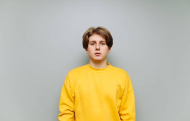 Sarı kazaklı yakışıklı genç adam gri arka planda izole edilmiş ve kameraya ciddi şekilde bakıyor..