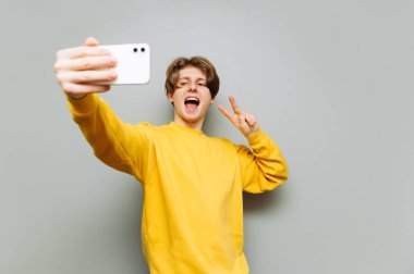 Parlak günlük giysiler içindeki mutlu adam gri duvar arkasındaki akıllı telefondan selfie çekiyor ve barış jesti gösteriyor. İzole edilmiş.