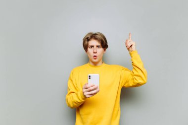Sarı günlük kıyafetli şaşkın bir adam gri bir arka planda elinde akıllı bir telefonla kameraya bakıyor ve şok olmuş bir yüzle baş parmağını gösteriyor..