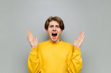 Şok olmuş adam gri arka planda duruyor ve kameraya çığlıklarla bakıyor ve ellerini kaldırıyor. Gri bir duvarın arkasındaki genç adam şaşırdı..