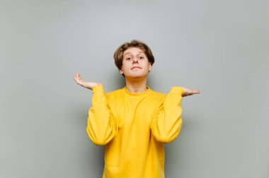 Sıradan giyinmiş yakışıklı bir adam kameraya bakar ve elleri ile bir karışıklık jesti gösterir. Gri bir arkaplanda izole edilmiş, komik bir yüzle kameraya bakar..