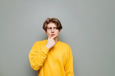 Sarı kazaklı genç adam gri bir arka planda duruyor ve düşünceli bir yüzle fotokopi odasına bakıyor. İzole edilmiş.