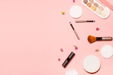Göz kamaştırıcı makyaj malzemeleriyle gösterişli düz çekim: parlak göz farı, mat ruj, fırçalar ve kurutulmuş çiçeklerle birlikte pastel pembe arka planda pamuk pedler..