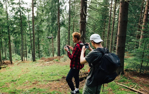 Two men create a video concentrate on a hike, a man shoots on camera as a friend flies a drone in a beautiful mountain forest. Create travel content from drones and cameras on the go. Backstage.