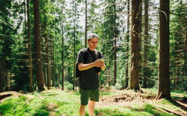 Gündelik giysiler içinde yakışıklı bir genç yaz günü sırtında bir sırt çantasıyla ormanda duruyor ve ciddi bir yüzle akıllı telefon kullanıyor..