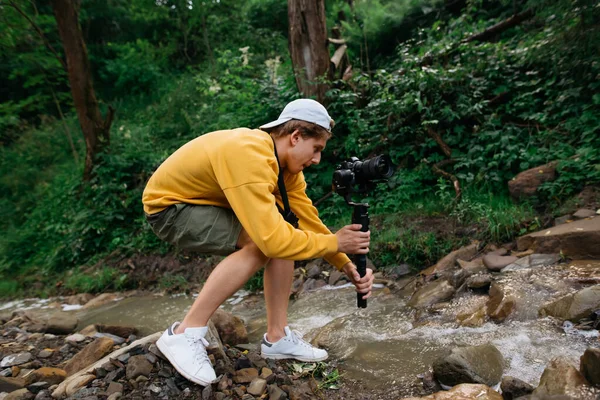 Young male cameraman with a camera on a stabilizer in his hands shoots a video of nature on a hike. Guy stands with a camera on a gimbal near a mountain stream and makes content.