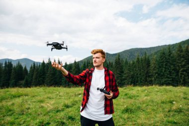 Kırmızı gömlekli yakışıklı genç adam, güzel bir manzaranın arka planında uzaktan kumandalı bir dron kontrolüyle bir dağ çayırında duruyor. Adam dağlarda İHA uçuruyor..
