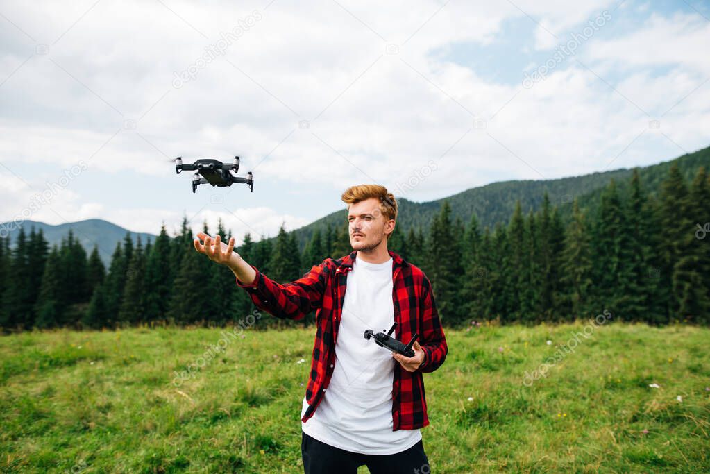 Handsome young man in a red shirt stands on a mountain meadow on a drone controls with a remote control on the background of a beautiful landscape. Guy flies a drone in the mountains.