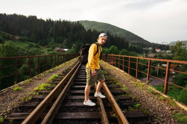 Turuncu kazaklı bir adam, dağlardaki bir demiryolu köprüsünde duruyor ve kameraya bakıyor. Genç erkek turist dağlarda viyadük üzerinde yürüyor.