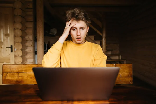 Anxious guy working on a laptop in a country house with a wooden interior. Nervous freelancer works at the computer on vacation.