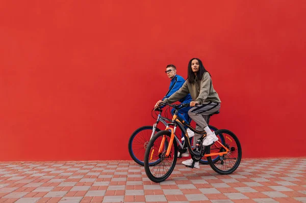 Photo of stylish man and woman riding a bicycle on the street on a background of a red wall. Stylish sports couple of cyclists walking down the street on the weekend
