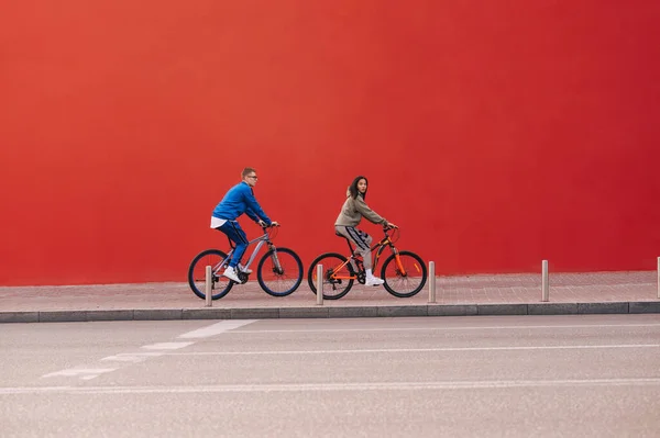 一个周末 一对时髦的夫妇骑着自行车 骑在红墙的背景下在城市里穿梭 红墙背景下两个骑自行车的人的简约照片 — 图库照片