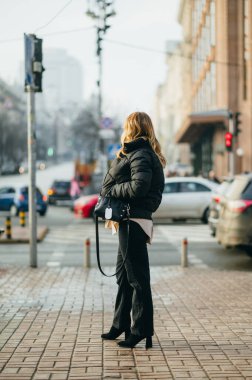 Sıradan giysiler içinde şık bir kadın şehrin arka planına karşı caddede duruyor ve yana bakıyor..