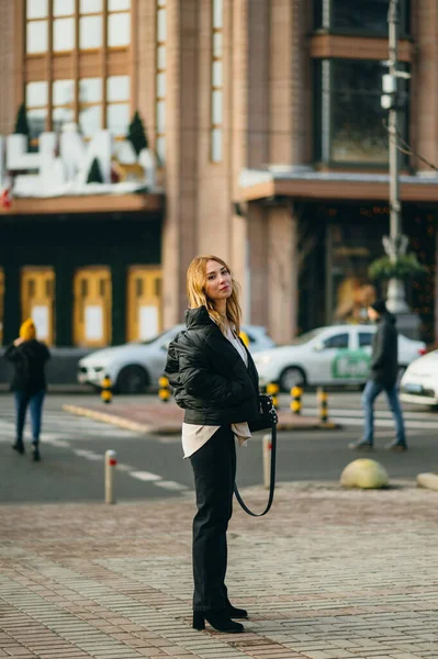 スタイリッシュなストリートウェアを着た魅力的な全長の女性が大都市の通りに立っており 彼女の顔に笑顔でカメラを見て ファッショナブルな流行の服を身に着けています — ストック写真