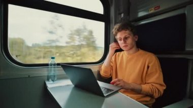 Kablosuz kulaklıklı genç adam tren kompartımanına biniyor ve ciddi yüzlü bir dizüstü bilgisayardan video izliyor. Trendeki masada dizüstü bilgisayarda çalışan serbest çalışan bir adam..