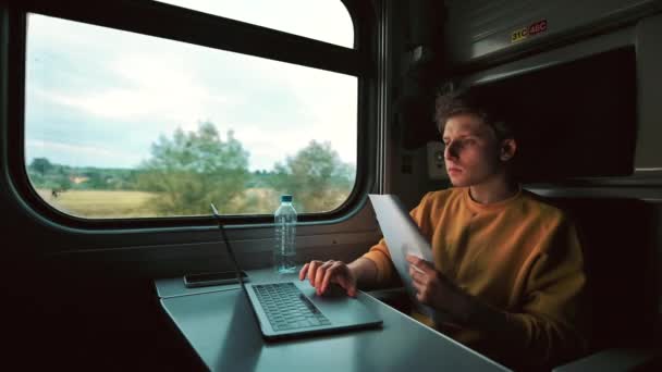 若いビジネスマンはノートパソコンを使用し 電車のコンパートメントで文書で動作します 学生の男は電車や研究で移動します — ストック動画