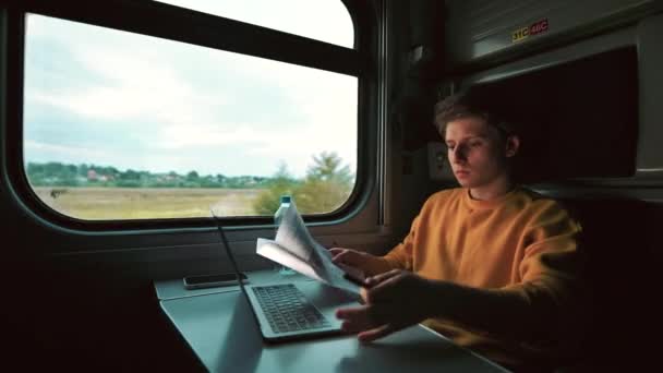 手にペンを持った青年が電車のコンパートメントにノートパソコンを持って座り 真剣な顔をした紙を読む フリーランサーは電車で旅行中にノートパソコンや新聞で働いています — ストック動画