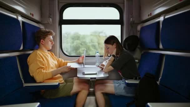 若い美しいカップルは電車で旅行し ノートパソコンやスマートフォンを使用しています 電車のコンパートメントの女性はスマートフォンを使用し 男はラップトップで動作します — ストック動画