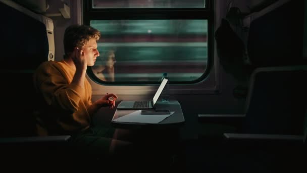 若い男は夕方に電車のコンパートメントに乗り 無線ヘッドフォンでラップトップを使用しています ラップトップでの移動 電車に乗ることで働くフリーランス — ストック動画