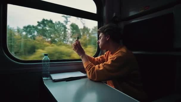 オレンジのセーターを着た若者が電車の中を歩き スマートフォンのカメラの窓から風景写真を撮る 鉄道旅行の概念 — ストック動画