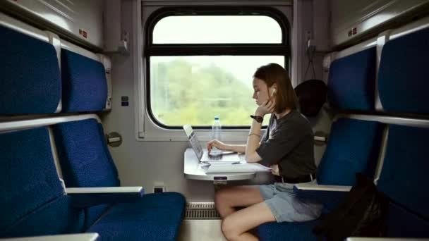 ビジネスカジュアルな服を着たかわいい女性は ラップトップ付きのテーブルの上のコンパートメント列車に座って 深刻な顔をした紙を読み取り ワイヤレスヘッドフォンで聞きます — ストック動画