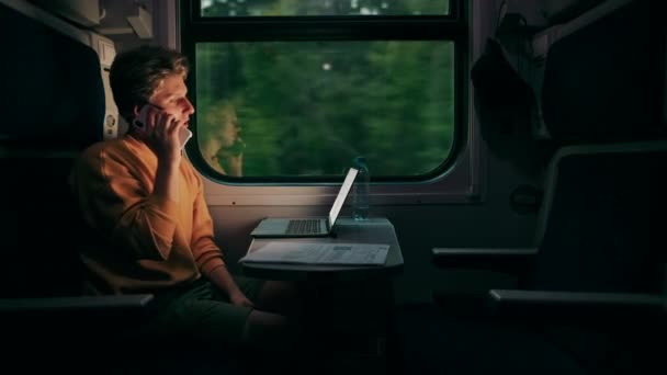 電車のコンパートメントのテーブルにノートパソコンを持って座って 旅行でオンラインで作業しながら電話をかける正の若い男 — ストック動画