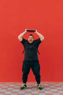 Siyah elbiseli komik şişman adam elinde dizüstü bilgisayarla siyah bir duvarın arkasında duruyor ve kameraya bakıp komik bir yüz ifadesi takınıyor..