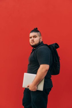 Siyah elbiseli yakışıklı ve kilolu bir adam kırmızı bir arka planda elinde dizüstü bilgisayarla ve sırtında bir sırt çantasıyla kameraya bakıyor.