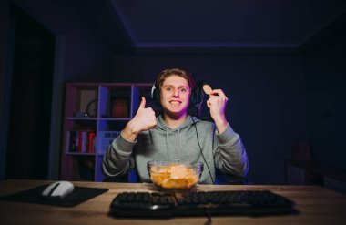 Kafasında kulaklık ve günlük kıyafetlerle komik bir adam geceleri elinde tabak ve çiplerle bilgisayara oturur ve yüzünde gülümsemeyle parmağını kaldırır..