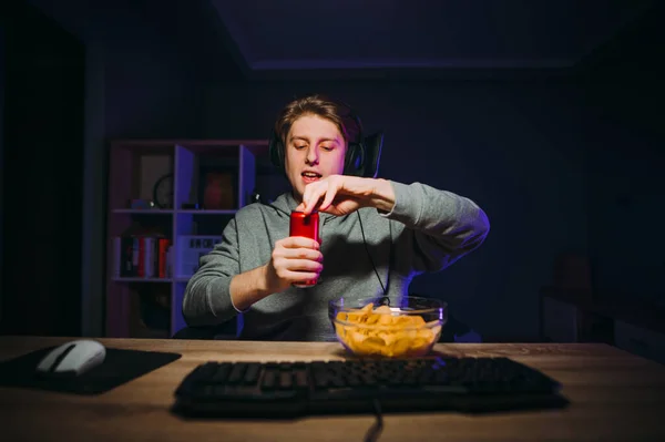 Guy in casual clothes and headphones on his head eats chips and cola in the bedroom at the computer at night, opens a can of drink with a smile on his face.