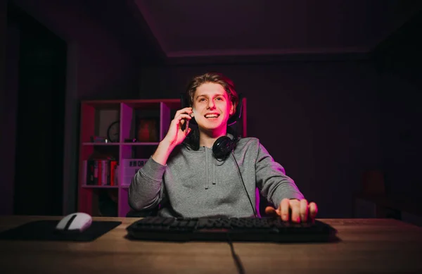 Handsome happy guy in a headset around his neck sits at a table at a computer in a room with a purple light and calls on the phone with a smile on his face.