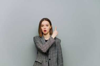 Gri arka planda duran ve şok olmuş yüzüyle baş parmaklarını gösteren ceketli kadın çalışanın portresi. Şok olmuş iş kadını boş alanda parmağını gösteriyor.
