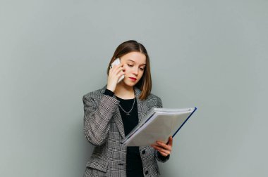 Gri bir arka planda duran ve akıllı telefondan yapılan aramalarda ciddi bir yüzle dokümanları okuyan şık giysili genç bir ofis çalışanı. Bayan iş yapıyor, izole..