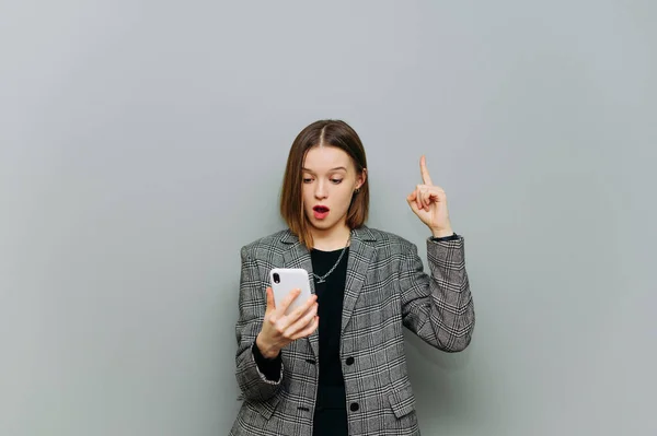 Shocked woman in smart casual clothes enjoys smartphone on gray background with surprised face and shows thumbs up on copy space.