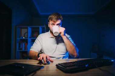 Beyaz tişörtlü, uykulu bir adam iş yerinde oturur ve bilgisayarda çalışır, fincandan kahve içer ve bitkin bir şekilde ekrana bakar..