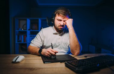 Kulaklıklı, üzgün bir adam geceleri evde grafik tabletindeki bir bilgisayarda çalışıyor ve gözlerini yorucu bir şekilde kapatıyor. Uykulu yorgun rötuşçu geceleri bilgisayar başında çalışır..