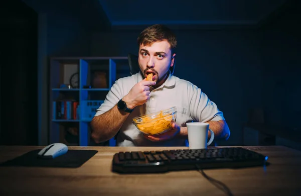 Hungry bearded man works and the internet at night at the computer and eats chips from a plate, looking at screen with a serious face.Night snack of a freelancer while working at home on the Internet.