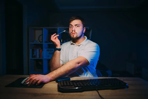 Surprised young man works at night on the computer and smokes an electronic cigarette, looking into the camera with a shocked face. Night work freelance, evaporation of tobacco.