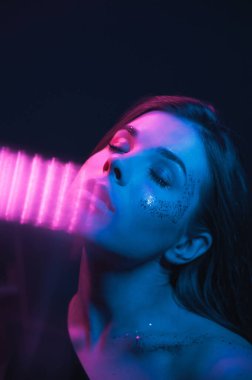 Mor ve mavi ışıkta parlak makyajlı, gözleri kapalı kameraya poz veren bir kadının moda gece portresi. Dikey.